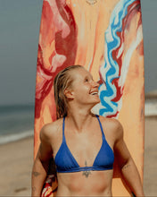 Load image into Gallery viewer, Woman in a blue surf bikini holding a colorful surfboard on a beach
